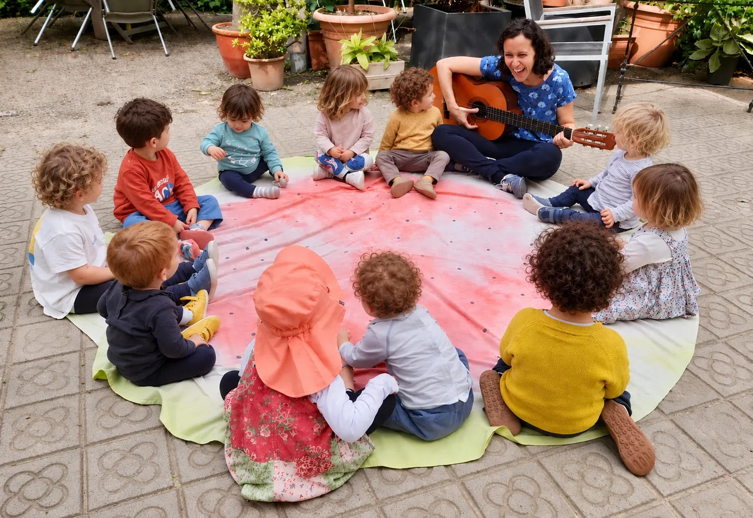 Infants asseguts en cercle sobre una manta al jardí mentre una acompanyant toca la guitarra