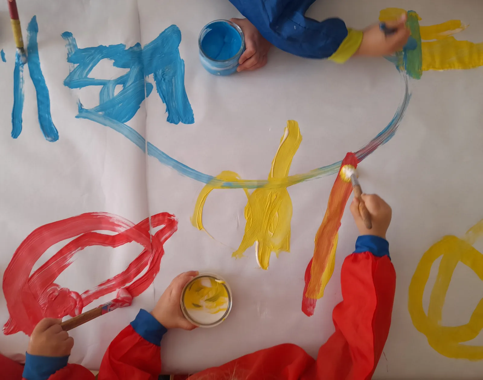 Mans d’infants pintant amb pintura de dits blava, groga i vermella sobre un paper gran